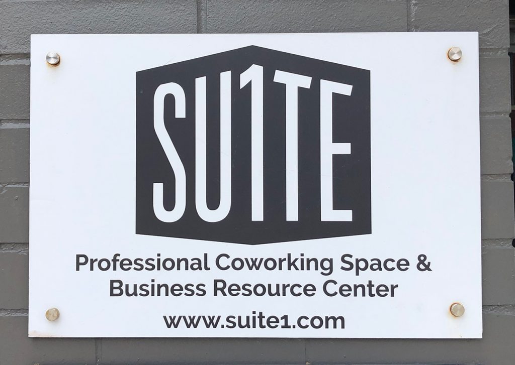 A white sign mounted with silver pegs against a gray-painted brick wall. On the sign is Suite 1 Logo | A black polygon on white ground with white letters that say SU1TE in a sans-serif font.