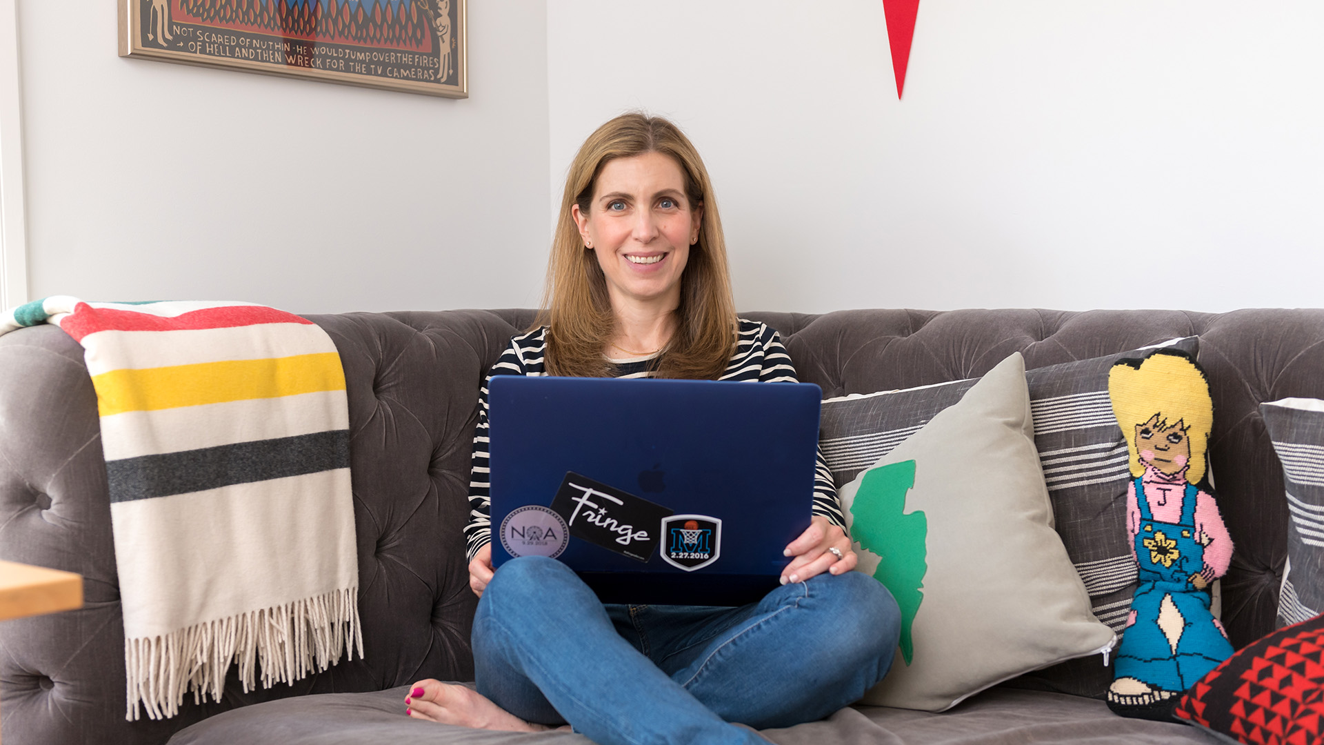 Smiling woman sits cross-legged on a gray couch, holding a laptop.