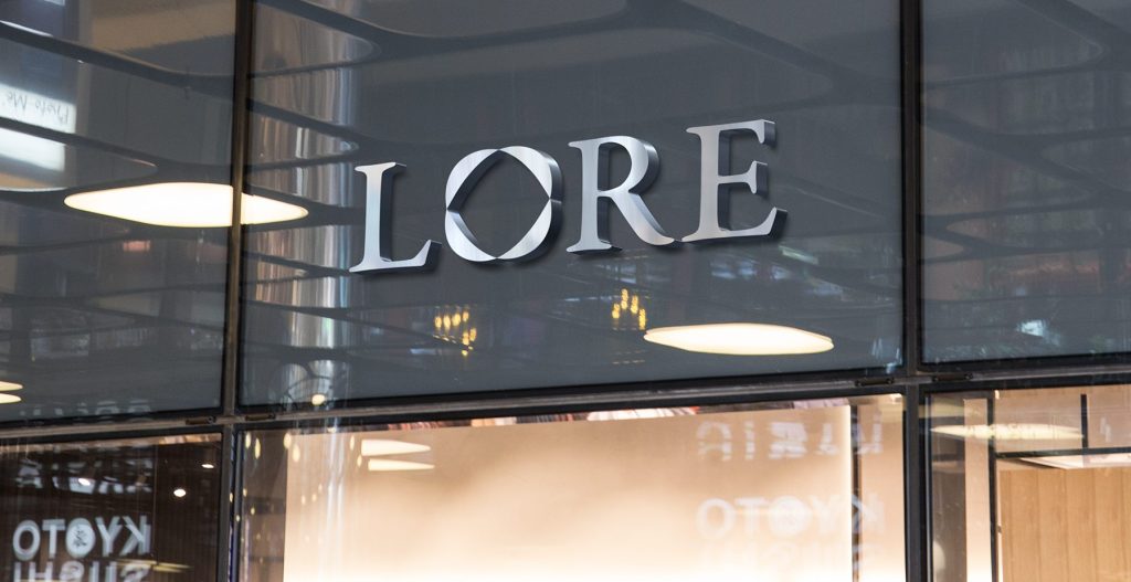 A photo mockup of the Lore logo rendered in silver metal letters mounted on a window of a modern glass storefront.