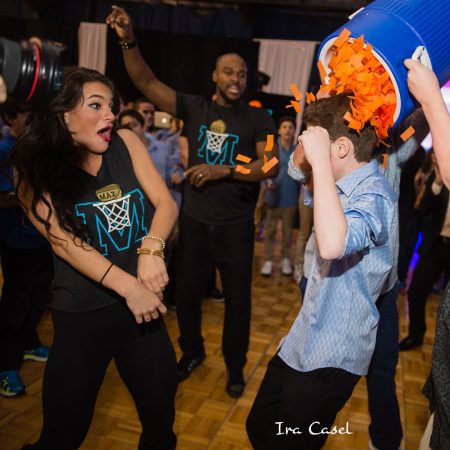 Female and male dancer wearing M basketball logo t-shirts, reacting to boy getting orange paper dumped on his head.